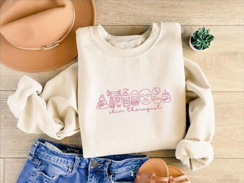 Puede incluir: Sudadera color crema con cuello redondo y mangas largas. La parte delantera presenta un dibujo lineal rosa de productos para el cuidado de la piel y las palabras "skin therapist". Un sombrero marr&oacute;n y unos vaqueros azules tambi&eacute;n est&aacute;n en la imagen.