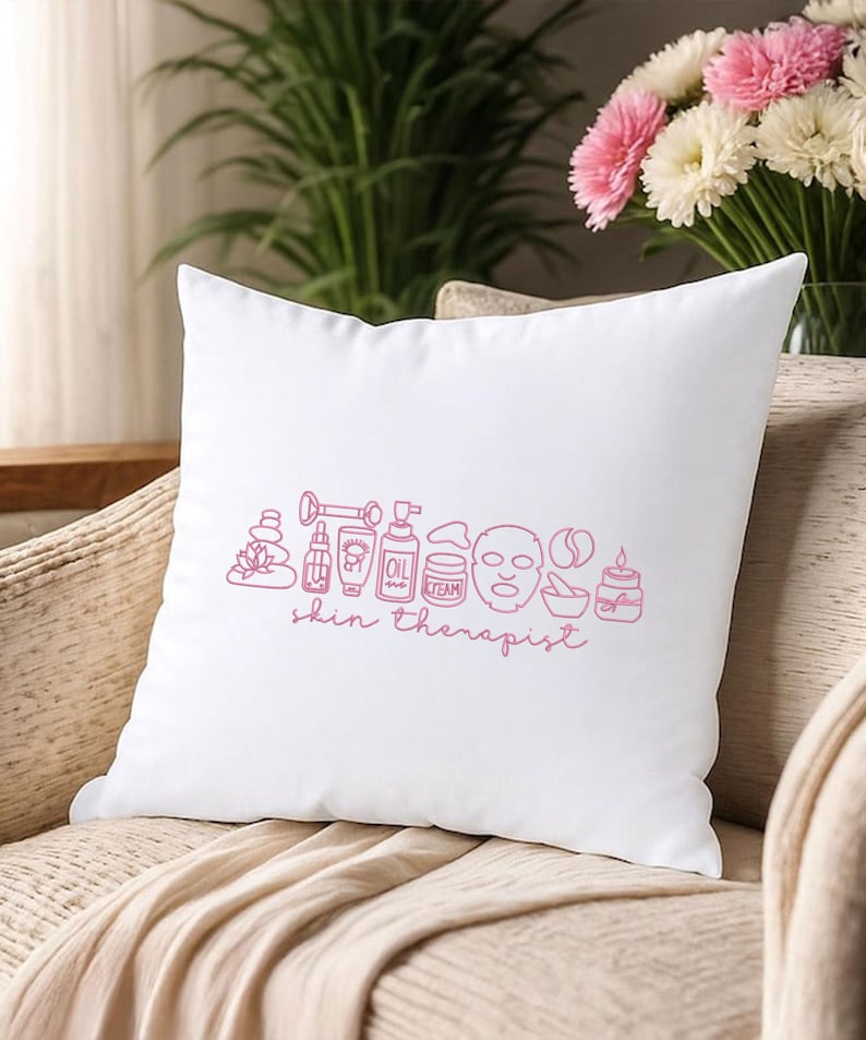 Puede incluir: Coj&iacute;n cuadrado blanco con un dibujo lineal rosa de art&iacute;culos de spa y cuidado de la piel, incluyendo una mascarilla facial y un frasco de aceite. Las palabras "skin therapist" est&aacute;n escritas en rosa debajo. El coj&iacute;n est&aacute; sobre una silla beige.
