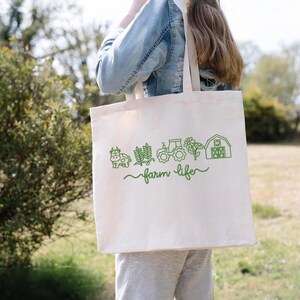 May include: A cream-coloured tote bag featuring a green farm-themed design. The design includes a cow, wheat, a tractor, a flower, and a barn, with the words "farm life" in a script font. The bag is being carried.