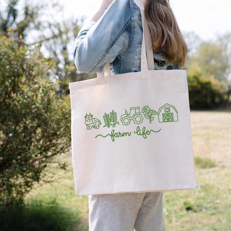 May include: A cream-coloured tote bag featuring a green farm-themed design. The design includes a cow, wheat, a tractor, a flower, and a barn, with the words "farm life" in a script font. The bag is being carried.