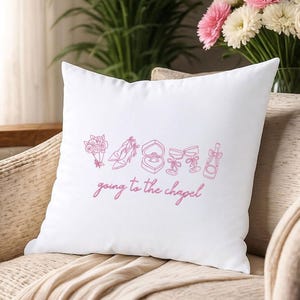 May include: White square pillow with pink line art of wedding-related icons, including a bouquet, shoes, ring, and champagne. The phrase "going to the chapel" is written in cursive below the icons. The pillow is on a beige chair.