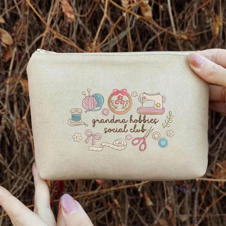 May include: A beige canvas pouch with a zipper closure. The pouch features a graphic of sewing and knitting tools, including yarn, a sewing machine, and scissors, with the text "grandma hobbies social club."