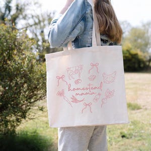 May include: A cream-colored tote bag with a pink illustration of farm animals and produce. The words "homestead mama" are written in cursive. The bag has two beige straps.