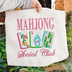 Może przedstawiać: Biała torba z r&oacute;żowym haftowanym napisem "MAHJONG Social Club". Projekt przedstawia trzy haftowane kafelki do Mahjonga w kolorze r&oacute;żowym, zielonym i niebieskim. Torba jest trzymana przez osobę.