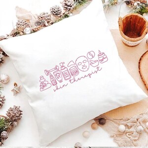 Puede incluir: Coj&iacute;n cuadrado blanco con un dibujo lineal rosa de productos para el cuidado de la piel y la frase "skin therapist". El dise&ntilde;o incluye una mascarilla facial, botellas de aceite y una vela. El coj&iacute;n est&aacute; sobre una superficie clara con pi&ntilde;as y un peque&ntilde;o cubo de madera.