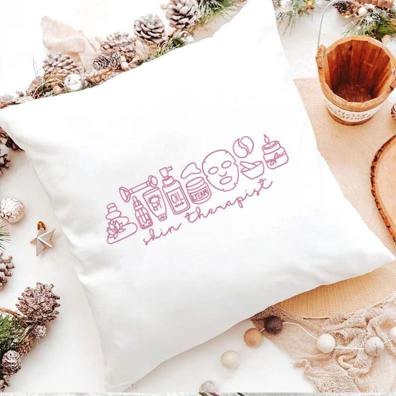 Puede incluir: Coj&iacute;n cuadrado blanco con un dibujo lineal rosa de productos para el cuidado de la piel y la frase "skin therapist". El dise&ntilde;o incluye una mascarilla facial, botellas de aceite y una vela. El coj&iacute;n est&aacute; sobre una superficie clara con pi&ntilde;as y un peque&ntilde;o cubo de madera.