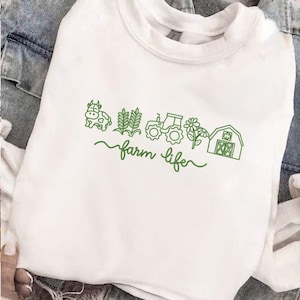 May include: White sweatshirt with a green outline design featuring a cow, wheat, tractor, flower, and barn. The words "farm life" are written in a cursive font below the illustrations. The sweatshirt is made of a soft material.