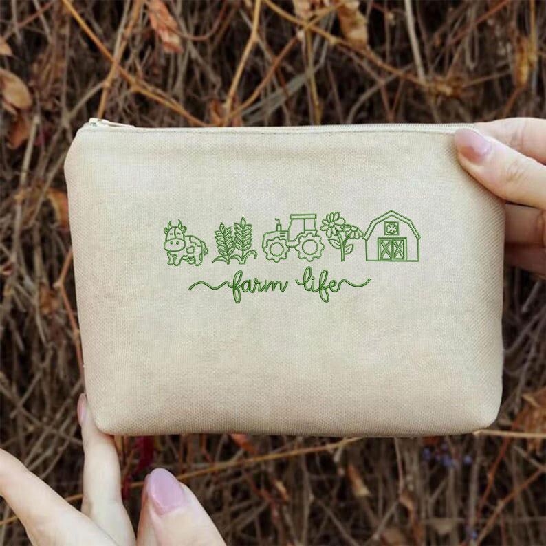 May include: A beige canvas pouch with a zip closure. The pouch features a green graphic of a cow, wheat, tractor, flower, and barn, with the words "farm life" in a script font. The pouch is held by a person.