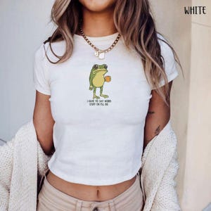 Peut inclure: Haut court blanc avec un dessin de grenouille tenant une tasse de caf&eacute;. La grenouille est verte et jaune, avec le texte "I HAVE TO SAY WEIRD STUFF OR I'LL DIE" en dessous. Le t-shirt a des manches courtes.