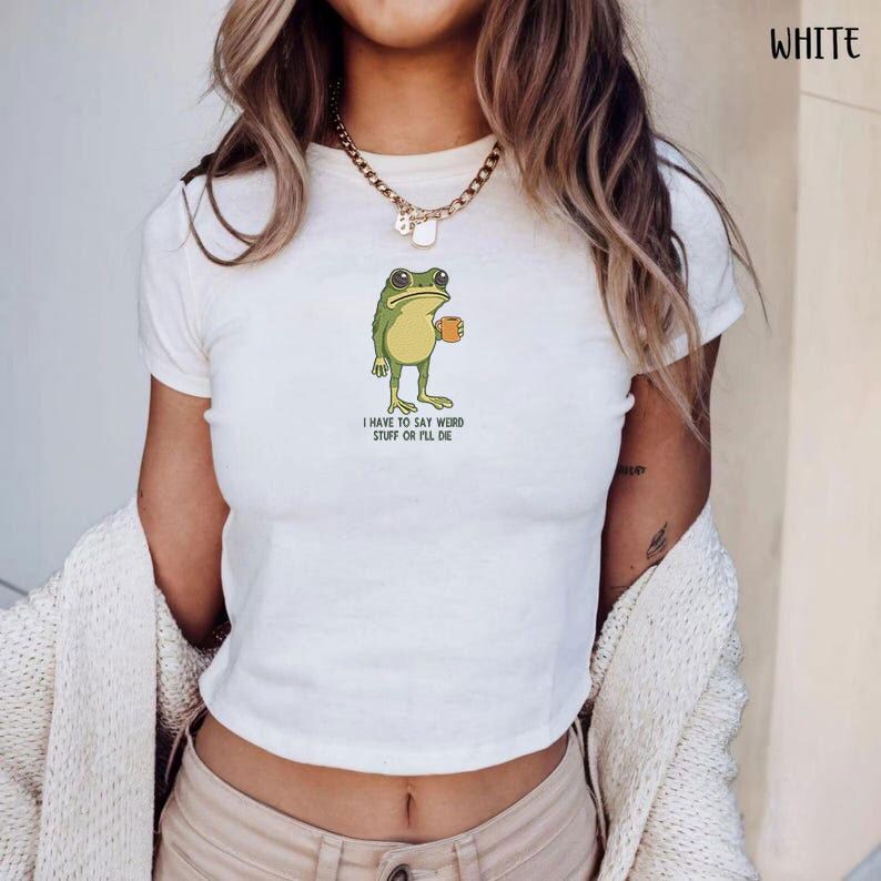 Peut inclure: Haut court blanc avec un dessin de grenouille tenant une tasse de caf&eacute;. La grenouille est verte et jaune, avec le texte "I HAVE TO SAY WEIRD STUFF OR I'LL DIE" en dessous. Le t-shirt a des manches courtes.