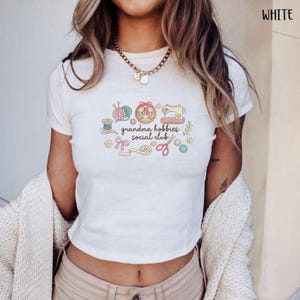 May include: White t-shirt with a colorful graphic that reads "grandma hobbies social club." The graphic includes images of yarn, a sewing machine, scissors, and other craft-related items. The shirt is short-sleeved and has a crew neck.