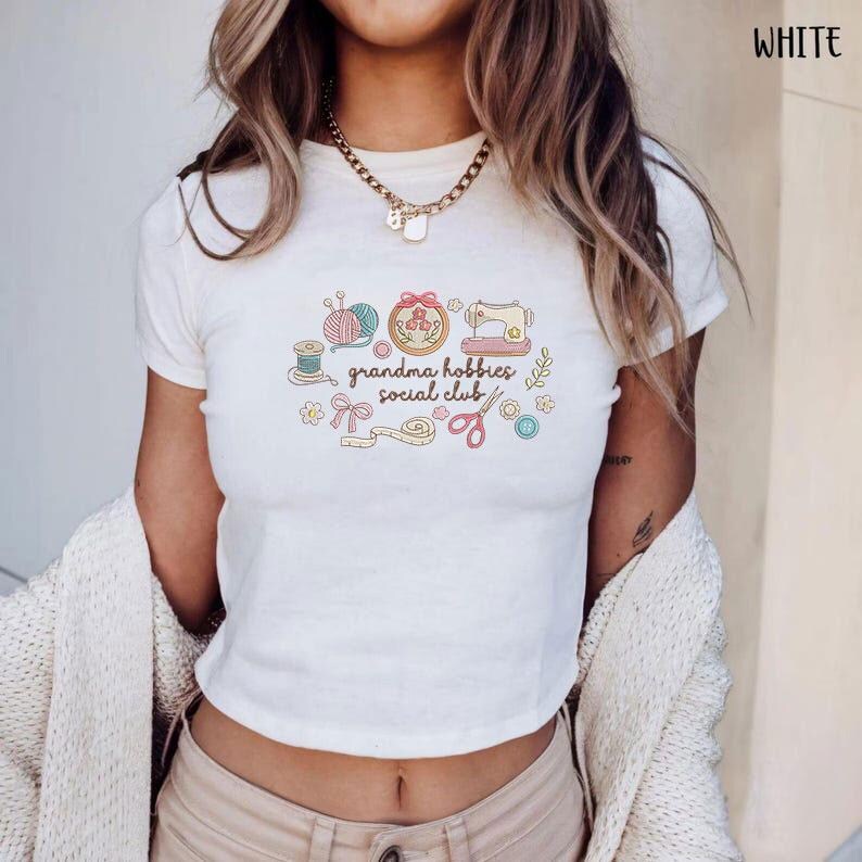 May include: White t-shirt with a colorful graphic that reads "grandma hobbies social club." The graphic includes images of yarn, a sewing machine, scissors, and other craft-related items. The shirt is short-sleeved and has a crew neck.