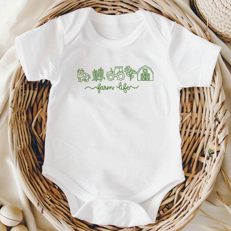 May include: White baby bodysuit featuring a green farm-themed illustration. The design includes a cow, wheat, a tractor, a sunflower, and a barn, with the words "farm life" in a cursive font. The bodysuit is displayed in a woven basket.