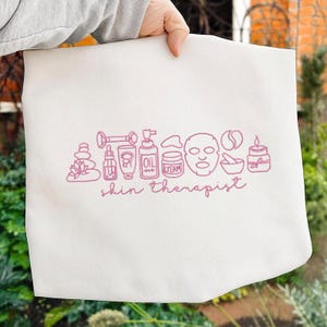 Puede incluir: Un bolso tote blanco con un dibujo lineal rosa de art&iacute;culos para el cuidado de la piel, incluyendo una mascarilla facial, un frasco de aceite y una vela. Las palabras "skin therapist" est&aacute;n escritas en rosa debajo.