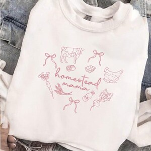 May include: White crewneck sweatshirt with a pink graphic. The design includes a cow, chicken, vegetables, and the words "homestead mama" in a cursive font. The sweatshirt is made of a soft material.