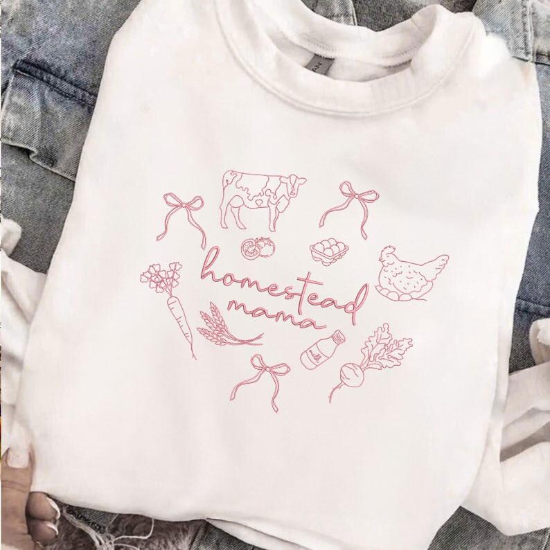 May include: White crewneck sweatshirt with a pink graphic. The design includes a cow, chicken, vegetables, and the words "homestead mama" in a cursive font. The sweatshirt is made of a soft material.