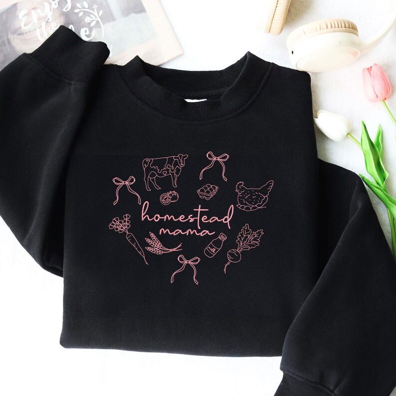 May include: Black crewneck sweatshirt with a pink graphic design. The design features a cow, chicken, milk bottle, and other farm-related illustrations, along with the words "homestead mama". The sweatshirt has long sleeves and a round neckline.