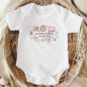 May include: White baby onesie with a colorful embroidered design. The design includes a sewing machine, yarn, embroidery hoop, scissors, and the text "grandma hobbies social club". The onesie is displayed in a woven basket.