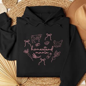 May include: Black hoodie with a pink graphic that says "homestead mama." The graphic includes illustrations of a cow, chicken, eggs, vegetables, and bows. The hoodie has a drawstring hood and a kangaroo pocket.
