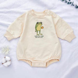 Peut inclure: Barboteuse b&eacute;b&eacute; cr&egrave;me &agrave; manches longues et col rond. La barboteuse pr&eacute;sente une grenouille de dessin anim&eacute; tenant une tasse, avec le texte "I HAVE TO SAY WEIRD STUFF OR I'LL DIE" imprim&eacute; en dessous.
