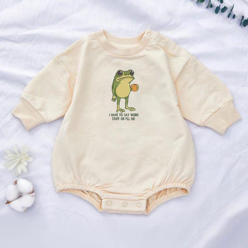 Peut inclure: Barboteuse b&eacute;b&eacute; cr&egrave;me &agrave; manches longues et col rond. La barboteuse pr&eacute;sente une grenouille de dessin anim&eacute; tenant une tasse, avec le texte "I HAVE TO SAY WEIRD STUFF OR I'LL DIE" imprim&eacute; en dessous.