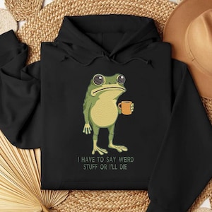Peut inclure: Sweat &agrave; capuche noir avec une grenouille de dessin anim&eacute; tenant une tasse. La grenouille est verte et jaune avec de grands yeux. Le texte "I HAVE TO SAY WEIRD STUFF OR I'LL DIE" est imprim&eacute; en dessous en blanc.