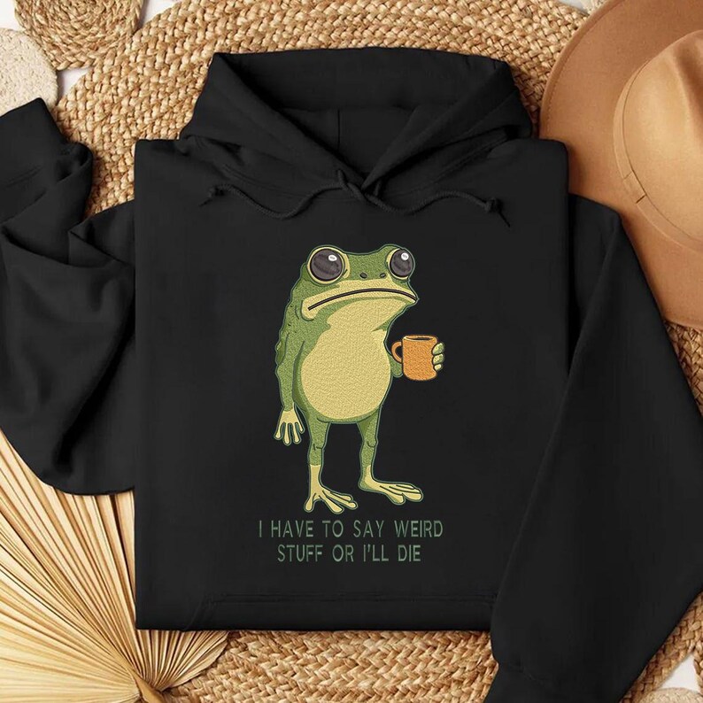 Peut inclure: Sweat &agrave; capuche noir avec une grenouille de dessin anim&eacute; tenant une tasse. La grenouille est verte et jaune avec de grands yeux. Le texte "I HAVE TO SAY WEIRD STUFF OR I'LL DIE" est imprim&eacute; en dessous en blanc.