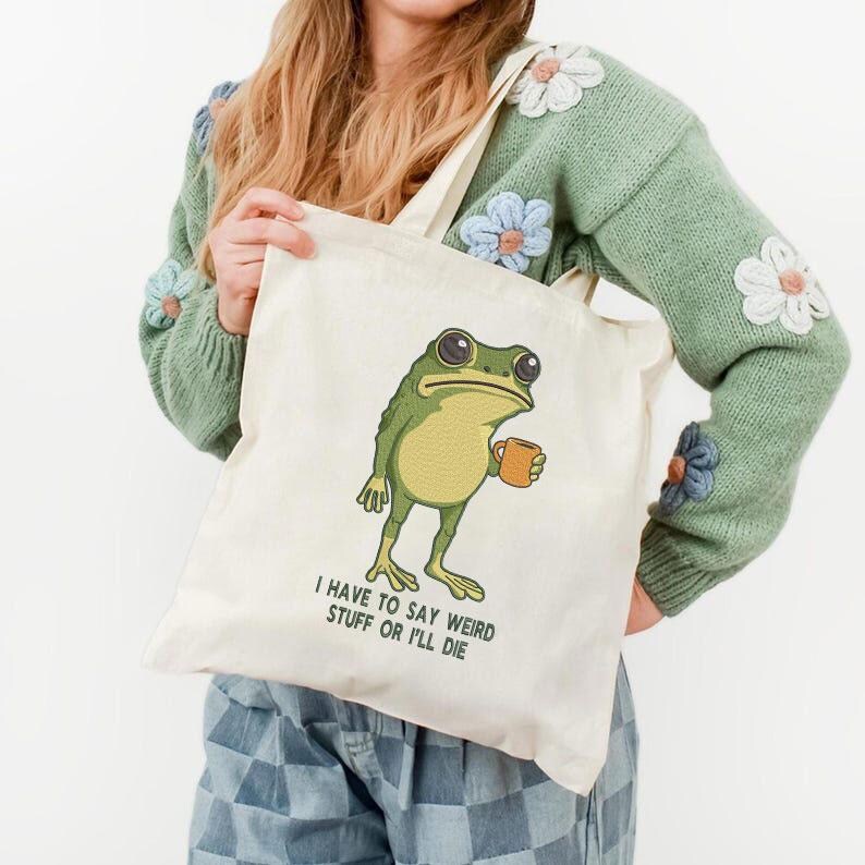 Peut inclure: Sac fourre-tout cr&egrave;me avec une grenouille de dessin anim&eacute; tenant une tasse. La grenouille est verte et jaune avec de grands yeux. Le texte sur le sac se lit "I HAVE TO SAY WEIRD STUFF OR I'LL DIE."