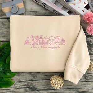 Puede incluir: Sudadera beige con un gr&aacute;fico rosa de art&iacute;culos para el cuidado de la piel y la frase "skin therapist". El gr&aacute;fico incluye un rodillo de jade, botellas, una mascarilla facial y una vela. La sudadera est&aacute; doblada.