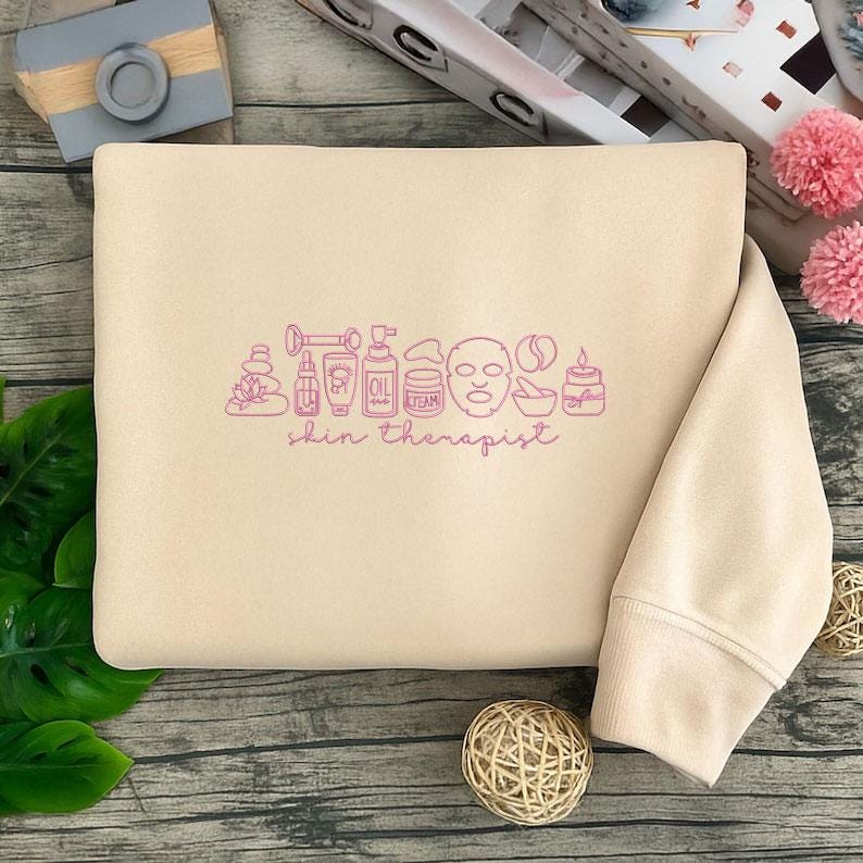 Puede incluir: Sudadera beige con un gr&aacute;fico rosa de art&iacute;culos para el cuidado de la piel y la frase "skin therapist". El gr&aacute;fico incluye un rodillo de jade, botellas, una mascarilla facial y una vela. La sudadera est&aacute; doblada.