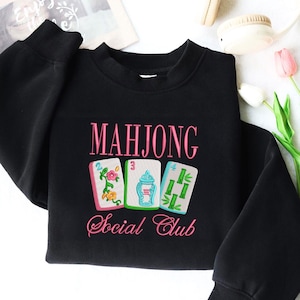 Może przedstawiać: Czarna bluza z haftowanym napisem "MAHJONG Social Club" w kolorze r&oacute;żowym. Poniżej znajdują się trzy haftowane kafelki do Mahjonga z kolorowymi wzorami. Bluza ma okrągły dekolt i długie rękawy.