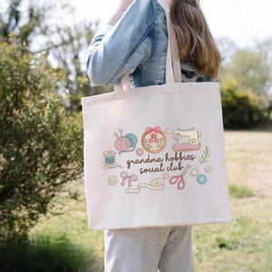 May include: Cream-colored tote bag featuring the text "grandma hobbies social club" with illustrations of sewing and knitting supplies. The design includes yarn, a sewing machine, and scissors.