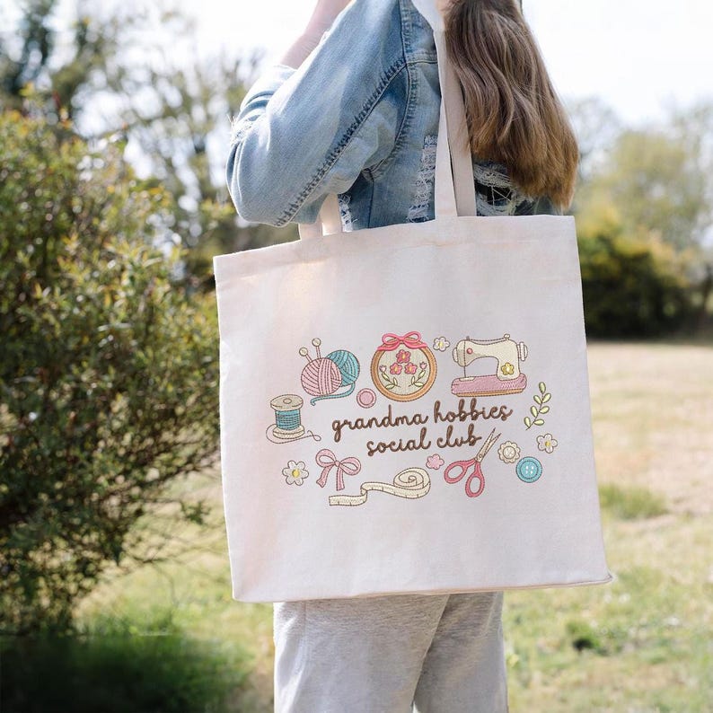 May include: Cream-colored tote bag featuring the text "grandma hobbies social club" with illustrations of sewing and knitting supplies. The design includes yarn, a sewing machine, and scissors.