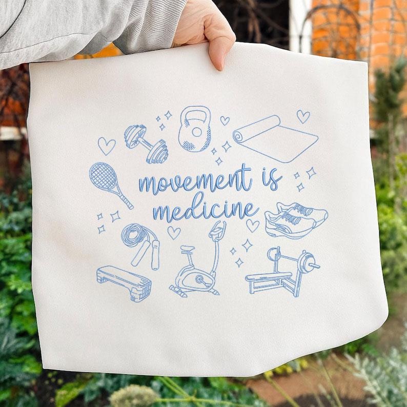 May include: White tote bag with blue line art of fitness equipment, including dumbbells, a kettlebell, and a yoga mat. The text "movement is medicine" is written in a script font. Hearts and stars are scattered around the illustrations.