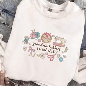 May include: White sweatshirt with a colorful embroidered design featuring crafting tools and the text "grandma hobbies social club". The design includes yarn, a sewing machine, scissors, and flowers.