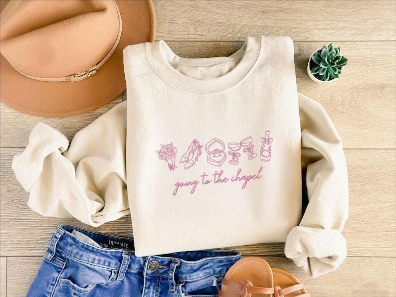 May include: Cream-colored sweatshirt with a crew neck, featuring a pink graphic of wedding-related icons and the phrase "going to the chapel." The sweatshirt is paired with blue denim shorts, a brown hat, and sandals.