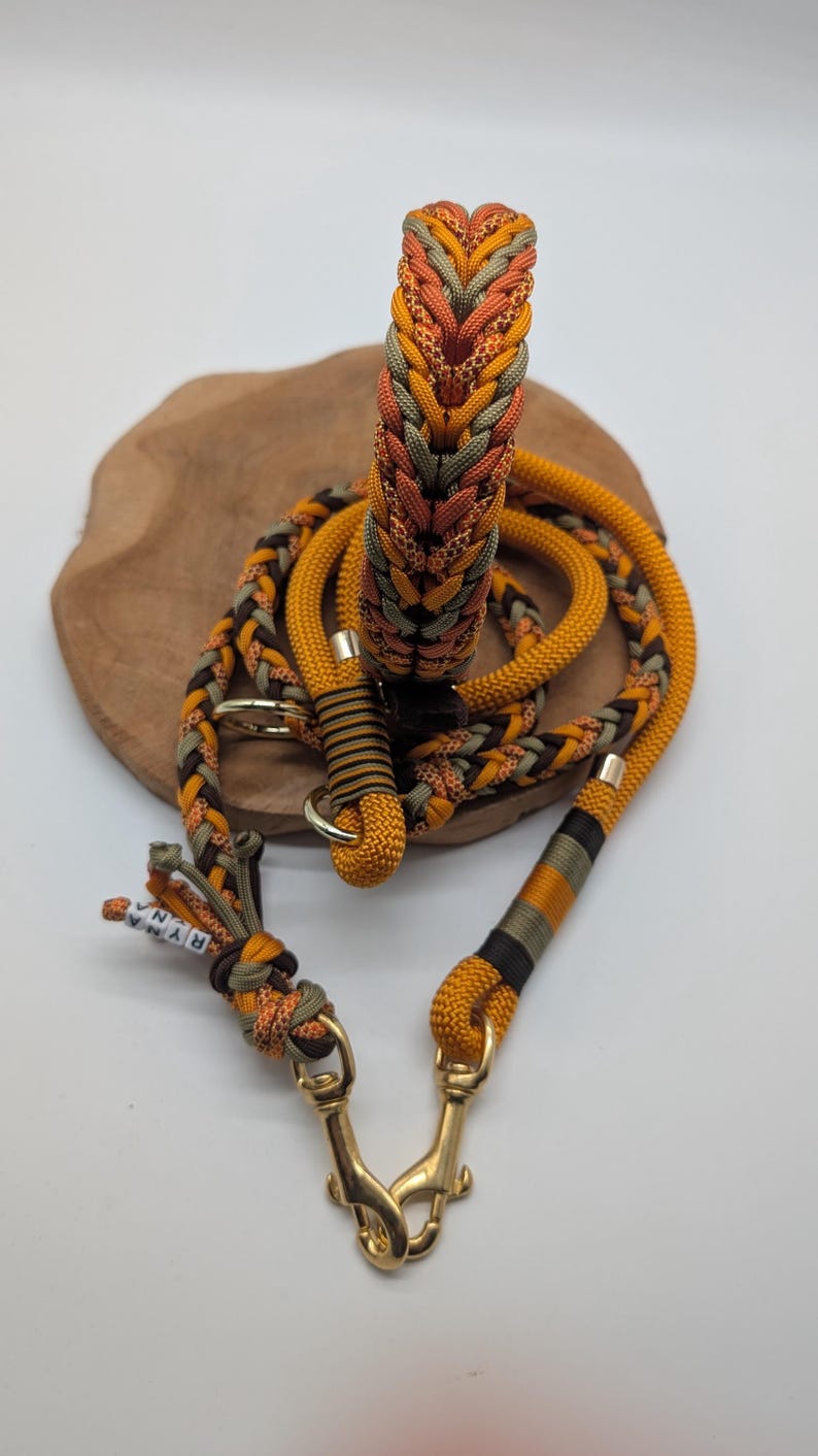 May include: A dog collar and leash set in shades of orange, brown, and green. The collar and leash are made of braided rope with gold-colored metal hardware. The set is displayed on a wooden surface.
