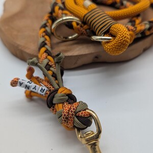 May include: A braided dog leash in shades of orange, brown, and olive green. The leash features a gold-colored clasp and a small section with the name "RYNA" spelled out in white beads. The leash is made of durable rope.
