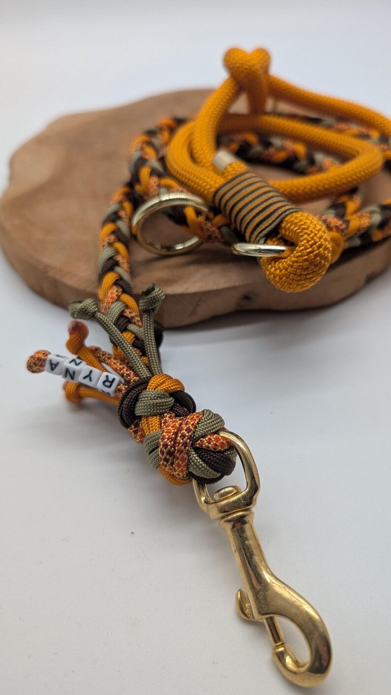 May include: A braided dog leash in shades of orange, brown, and olive green. The leash features a gold-colored clasp and a small section with the name "RYNA" spelled out in white beads. The leash is made of durable rope.