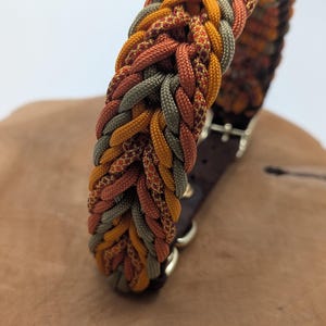 May include: Braided dog collar in shades of orange, yellow, and green. The collar features a brown leather backing and metal hardware. The woven design creates a textured appearance. A stylish accessory for pets.