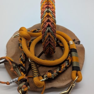 May include: A collection of dog accessories, including a braided collar and matching leash. The items feature a color scheme of orange, brown, and olive green, with gold-colored metal hardware. The leash has a clip for easy attachment.