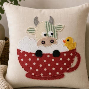 Peut inclure: Un coussin carré beige avec une applique en tissu représentant une vache aux oreilles rayées vertes et au museau beige, assise dans une tasse à thé rouge à pois blancs remplie de mousse, avec un petit canard en caoutchouc jaune sur le côté.