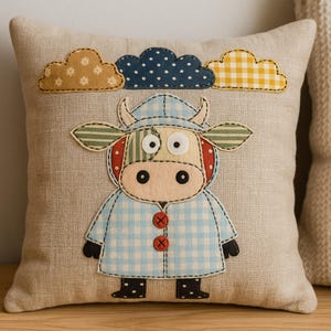 Puede incluir: Cojín cuadrado beige con un diseño de vaca aplicado. La vaca lleva un abrigo azul a cuadros y tiene una cara de retazos. Encima de la vaca hay tres nubes de tela en amarillo, azul y beige.
