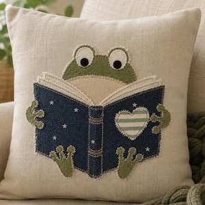 Peut inclure: Un coussin carré beige orné d'une grenouille verte en appliqué tenant un livre bleu foncé ouvert. La couverture du livre présente des motifs d'étoiles blanches et un cœur rayé. La grenouille a de grands yeux blancs et des mains palmées.