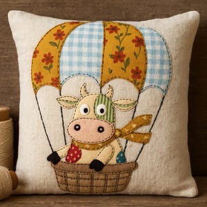 May include: Cream-colored square pillow featuring a whimsical cow design. The cow is in a basket suspended from a hot air balloon with a yellow floral and blue gingham pattern. The cow has a scarf and is made of various fabric patterns.