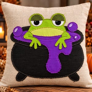 May include: A square beige throw pillow featuring a fabric appliqué of a bright green frog sitting inside a black cauldron filled with purple liquid. The frog has large eyes and the purple liquid spills over the side of the black pot.