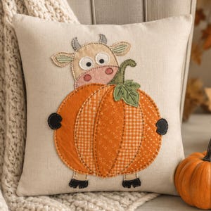 Puede incluir: Un cojín cuadrado de color beige con una aplicación de tela de una vaca que sostiene una gran calabaza naranja. La vaca tiene una cara de color tostado claro, ojos blancos y pezuñas negras. La calabaza está hecha de varias telas naranjas estampadas con una hoja verde.