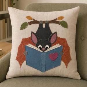 Puede incluir: Un cojín cuadrado de tela beige claro con una aplicación de fieltro de un murciélago negro colgado boca abajo de una rama marrón. El murciélago lee un libro azul decorado con un corazón rosa, flanqueado por alas naranjas y hojas verdes.