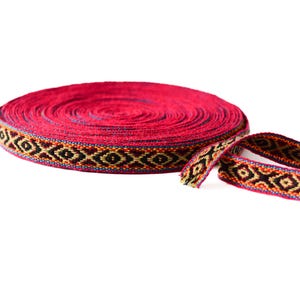 Può includere: Un rotolo di nastro tessuto rosso vibrante con un motivo geometrico nero, oro e arancione. Una sezione del nastro è srotolata, mostrando il design dettagliato. Questo nastro decorativo è ideale per l'artigianato e gli abbellimenti.