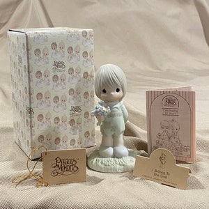 May include: A Precious Moments porcelain figurine of a young child holding flowers, with a matching box and informational cards. The figurine is light blue and white, with the words "I Belong To The Lord" on a card. The box has a pattern of cherubs.